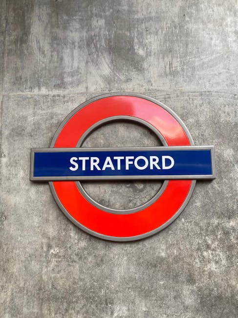 A close-up of a London Underground sign with a red circular border and a blue rectangular bar in the center displaying the word 'STRATFORD' in white uppercase letters, mounted on a textured concrete w