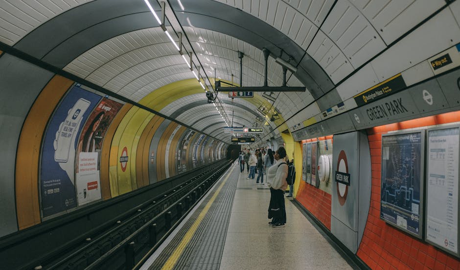 A view of the Green Park underground station platform with curved, multicolored walls in yellow, orange, and gray. Several passengers are standing on the platform, some holding bouquets of fresh flowe