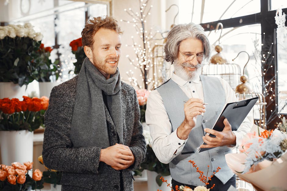 A professional florist from cockfostersflowers.co.uk, wearing a light grey vest and glasses, is holding a tablet and smiling while discussing floral arrangements with a male customer in a flower shop.