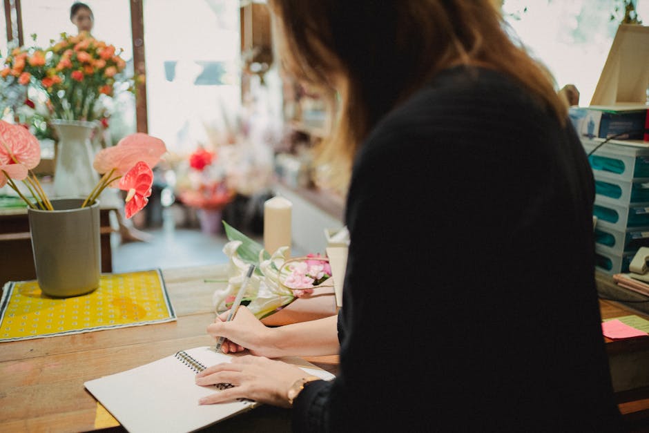 A florist in a flower shop is arranging a floral gift comprising pink roses, green leaves, and decorative filler flowers on a wooden table. The bouquet is wrapped in soft pink paper with a white ribbo