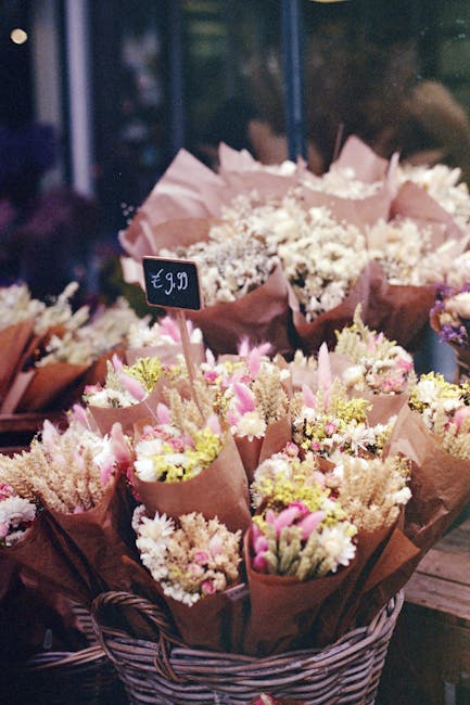 A display of multiple flower bouquets arranged in a woven basket, featuring soft pink, white, and yellow flowers wrapped in pink and brown paper. The bouquets include delicate carnations, tiny daisies