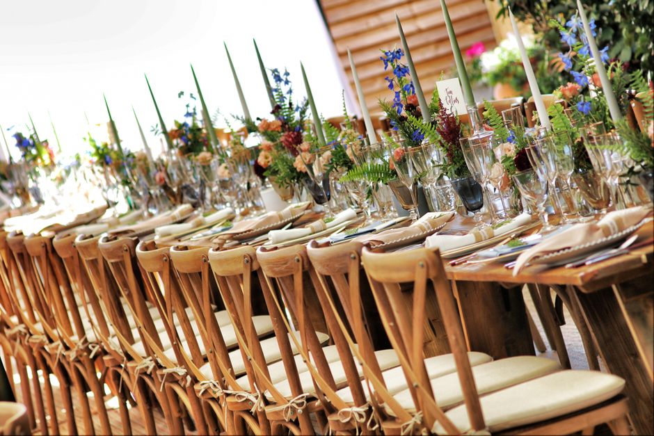 A long wooden dining table set for an event at Trent Park, arranged with elegant floral centerpiece arrangements featuring a mix of blue delphiniums, peach roses, pink carnations, and green ferns in c