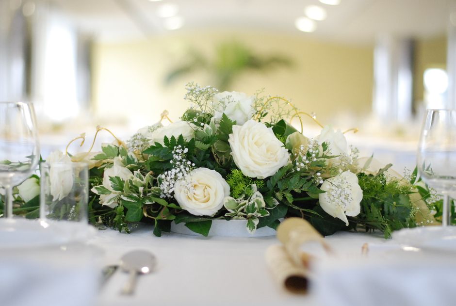 A floral centerpiece featuring white roses, calla lilies, and delicate baby’s breath, surrounded by lush green foliage including variegated leaves, arranged in a low, elongated form on a white table