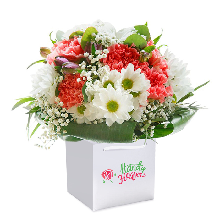 Elevated by an immaculate white square gift bag and sturdy ribbon handles, this resplendent floral arrangement captures the essence of Cockfosters charm. Clustered coral-pink and peach carnations, instantly recognisable by their delicately frilled edges, provide a plush, full-bodied centre, enriched by the drama of vividly red carnations interspersed for contrast. White daisies, all with striking lime-green centres, give a lively, playful accent and summon thoughts of open meadows near Oak Hill Park. Wispy clusters of gypsophila contribute a soft, ethereal quality, echoing mist in the gentle morning air. At the left, partially closed buds tinged in purple and blush insert a note of anticipation, promising beauty yet to be revealed. Surrounding the bouquet is a sumptuous layer of broad-leafed, glossy green foliage, creating a seamless, lush collar that structures and supports the blooms. The composition's rounded shape speaks to skilled arrangement techniques while maintaining an organic, naturally abundant appearance. Perfectly posed against a spotless white background, each nuance of tone and texture is laid bare, celebrating freshness, quality, and attention to detail expected from Cockfosters Flowers. This bouquet is equally at home as a luxurious birthday gift, a spontaneous delivery along Cockfosters Road, or as a centrepiece in a welcoming family home within the Cockfosters locality.